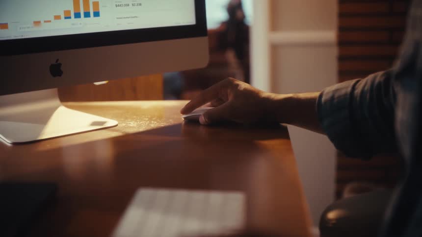 A person analyzes financial data on a computer in a warmly lit office, with a slow-motion focus pull to their hand using a mouse