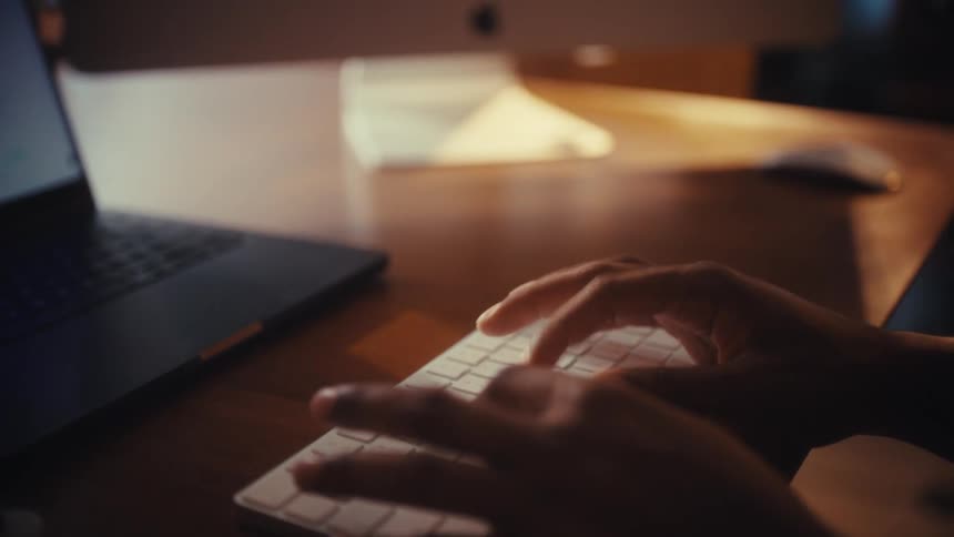 A cinematic close-up of a person's hands typing on a wireless keyboard in a warmly lit home office at night