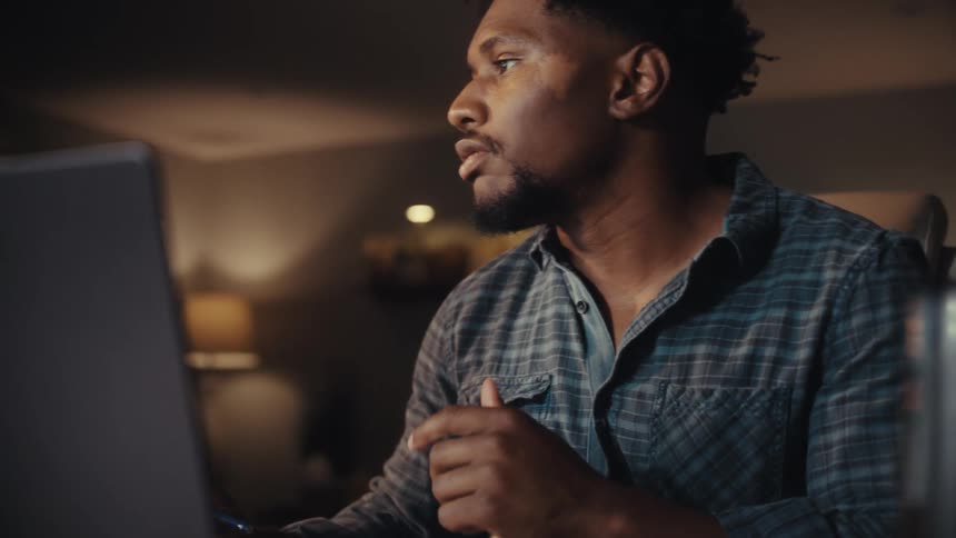 A thoughtful African American man works from home late at night, concentrating on his laptop screen in a dimly lit room