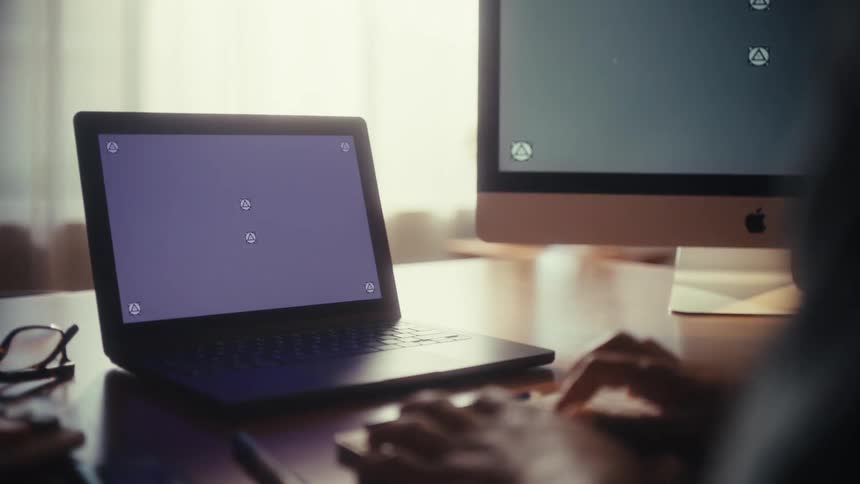 Over-the-shoulder shot of a person working at a desk with a laptop and desktop computer featuring blank screens for screen replacement