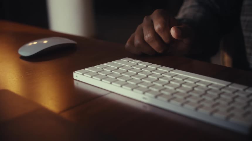 A person works in a dimly lit office, typing on a wireless keyboard before using a mouse on a warm wooden desk