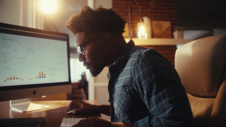 A slow dolly reveals a young professional man intently focused on analyzing data charts on his computer while working from his home office at night