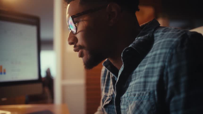A focused male professional wearing a plaid shirt and glasses intently studies business analytics on his desktop computer