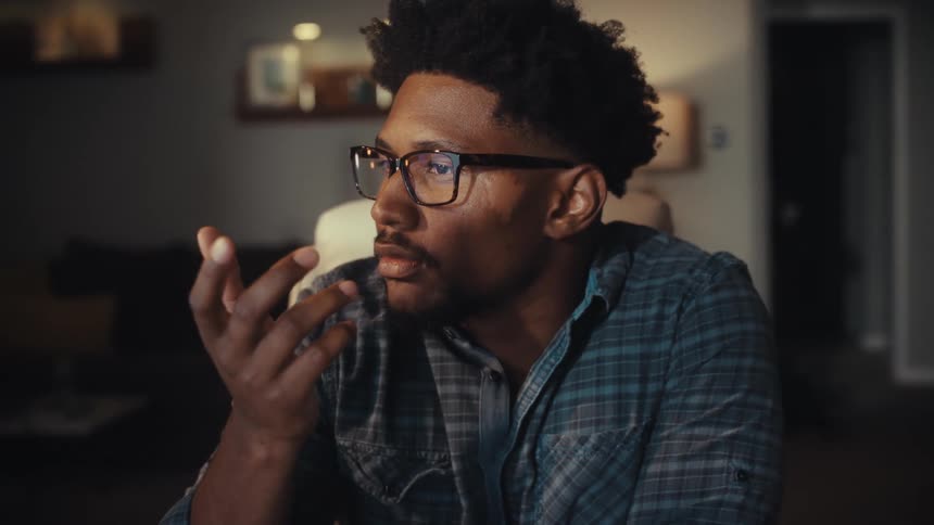 A thoughtful young Black man with glasses works from home, deeply focused on his computer screen