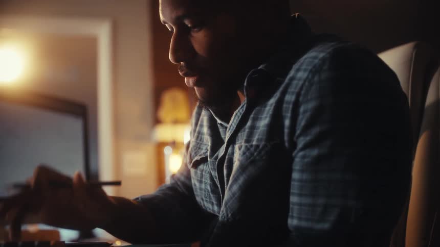 A focused African American man works late at night in a dimly lit room using a calculator and a laptop