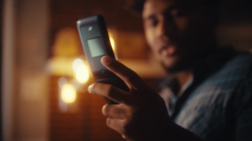 A man smiles while texting on a retro flip phone in a warmly lit room at night