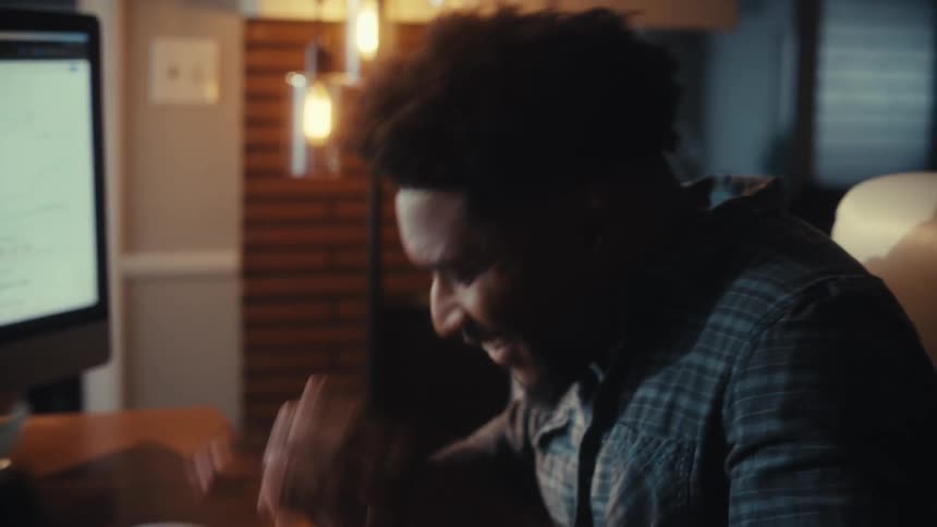 A distraught African American man panics while on the phone and looking at financial charts on his computer in a dimly lit home office