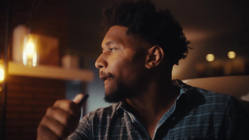 A focused young Black man in a plaid shirt works late, analyzing data on his computer in a dimly lit, warm office setting