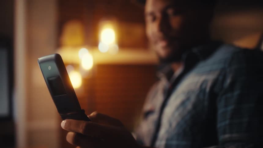 Close-up rack focus shot of an African American man smiling while using a retro flip phone in a cozy, warmly lit office at night