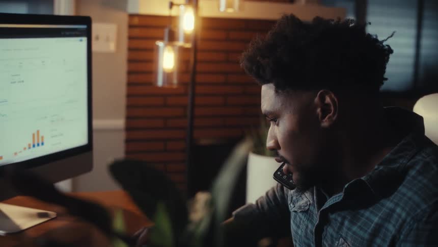 A focused young man multitasks by talking on the phone and analyzing business data on his computer while working late in his home office