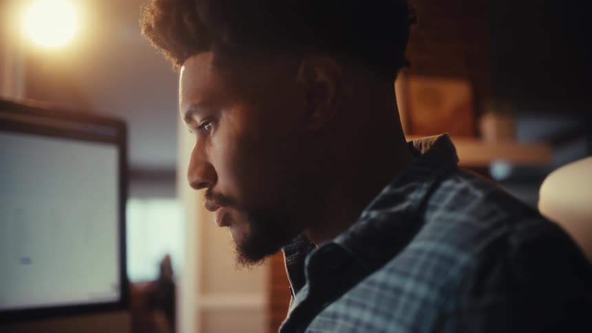 A focused young Black man works on his desktop computer in a modern home office with warm, cinematic lighting