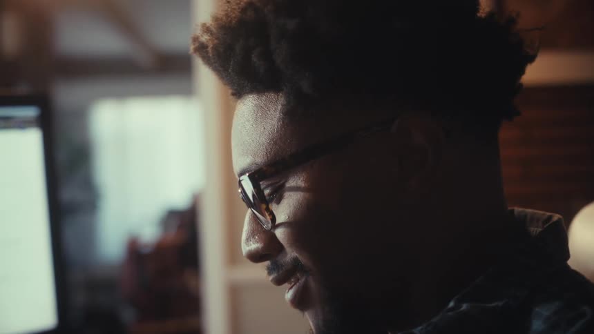 A young man with glasses smiles as he looks at data and charts on his computer while working from his warmly lit home office