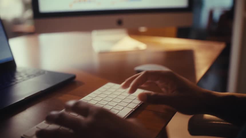 Slow-motion close-up of a person's hands typing on a wireless keyboard at a sunlit desk with computers in the background
