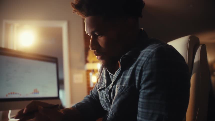 A young man concentrates while working from his home office late at night, using a calculator and analyzing data on his computer