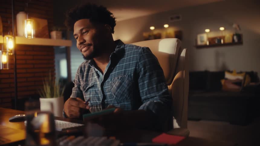 A young man works late in his warmly lit home office, talking on a video call and smiling as he writes a check