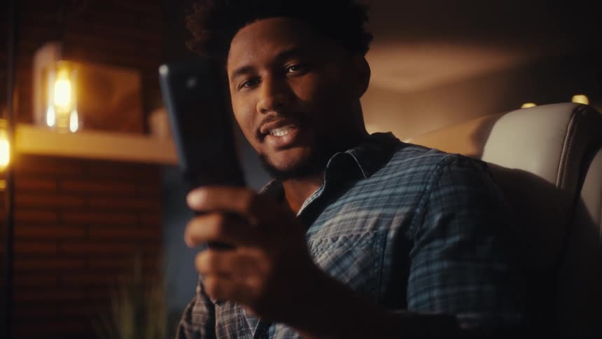 A relaxed young man smiles while scrolling on his smartphone in a modern, warmly lit room at night