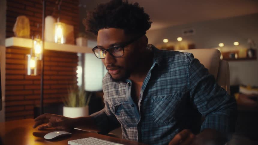 Slow zoom into a concentrated young man working on his computer setup in a stylish, dimly lit home office at night