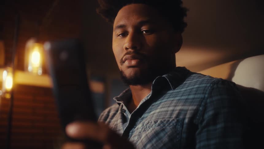 A handsome young Black man smiles while scrolling through his smartphone in a warmly lit room at night