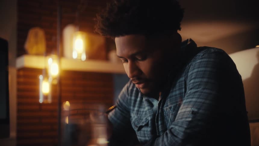 A focused young African American man thinks deeply while working at his computer in a warmly lit room at night