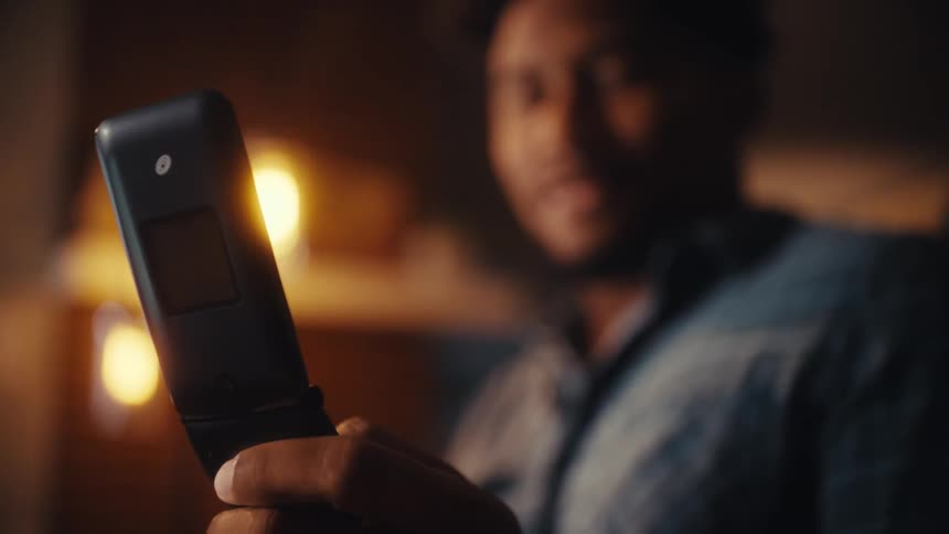 Close-up of a man smiling while holding a nostalgic flip phone in a warm, cozy room at night