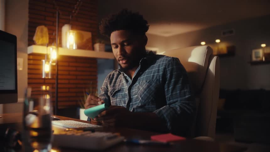 A focused young man works late at his home office desk, writing in a notepad while looking at his computer in a warmly lit room