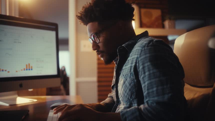A tired businessman works late from his home office, analyzing charts and data on his computer before taking off his glasses from stress