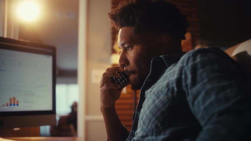 A concentrated businessman working late at his home office makes a serious phone call while reviewing charts on his computer
