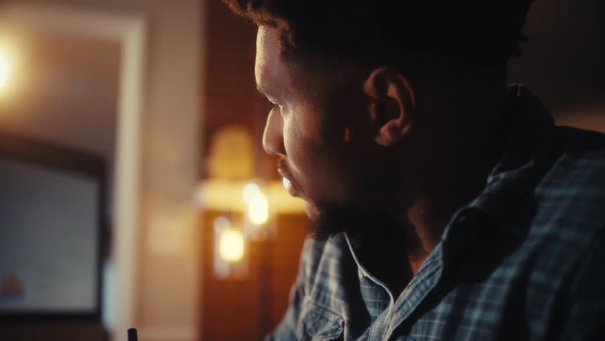 A focused young man in a plaid shirt works diligently, writing notes at his desk in a warmly lit home office at night