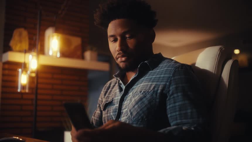 A focused young man working on his computer late at night in his dimly lit home office takes a break to use his smartphone