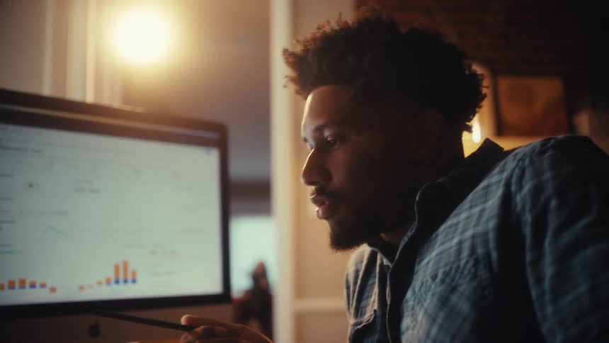 Cinematic close-up of a focused young African American man working late at night, analyzing data charts on his computer in a warmly lit home office