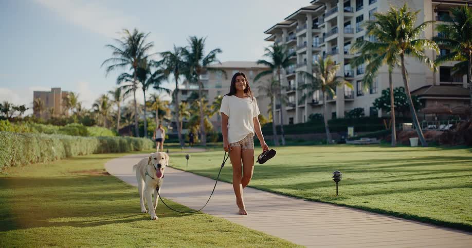A smiling woman walks barefoot with her golden retriever on a path through a tropical resort in slow motion