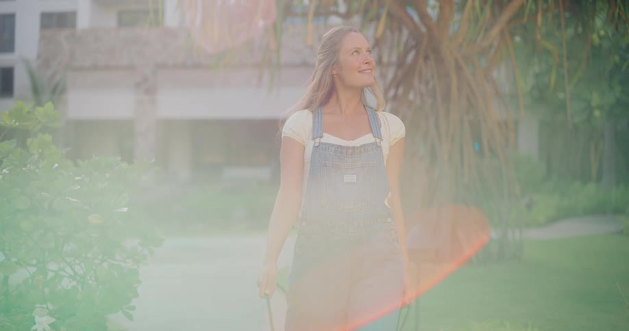 A happy young woman in overalls walks her golden retriever in slow motion on a path at a tropical resort with a beautiful sun flare