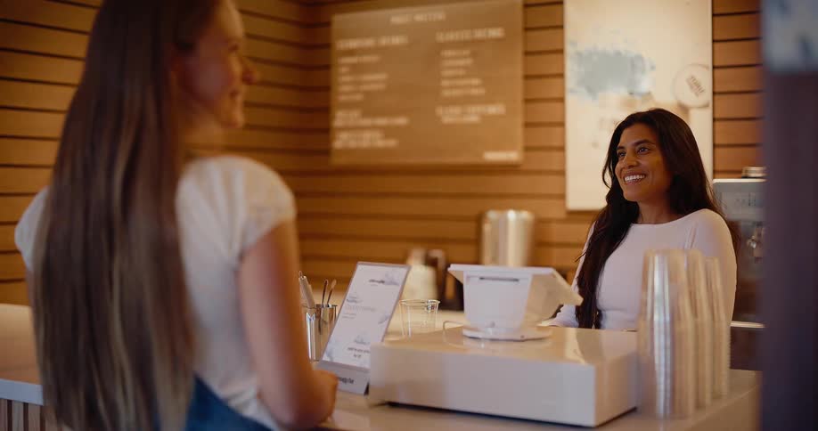 A friendly cashier with long dark hair smiles warmly as she greets a customer at the counter of a modern cafe