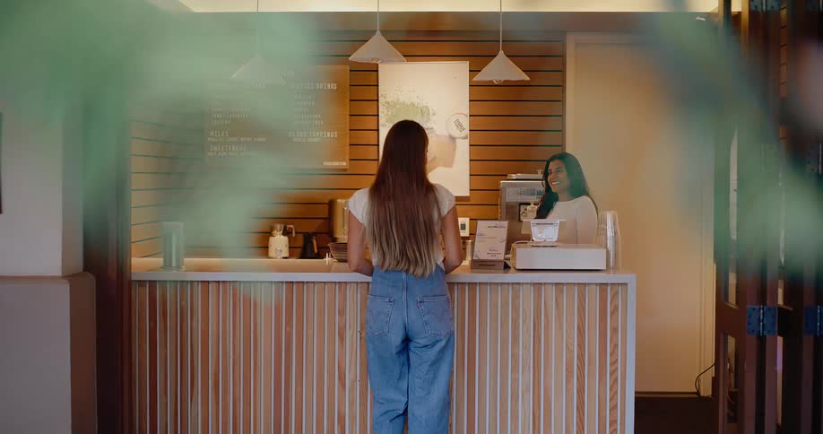 A young woman walks up to the counter of a modern cafe where a friendly barista waits to take her order