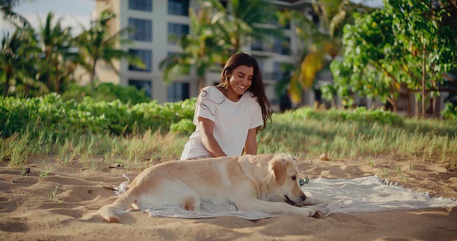 A smiling young woman pets her golden retriever as they relax together on a blanket at a sandy beach in slow motion