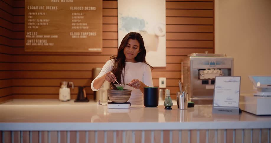 A woman carefully sifts green matcha powder into a bowl while preparing a traditional tea drink in a modern cafe