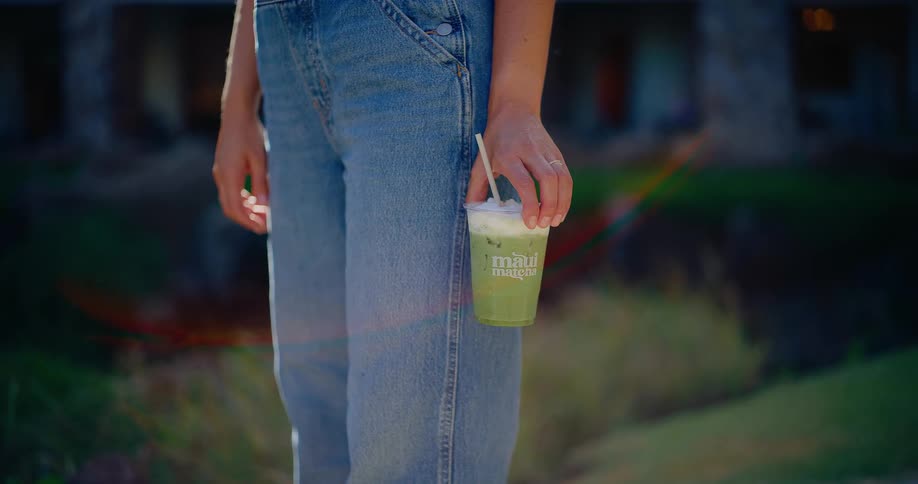 A woman wearing blue denim overalls holds a refreshing iced matcha latte in the afternoon sun