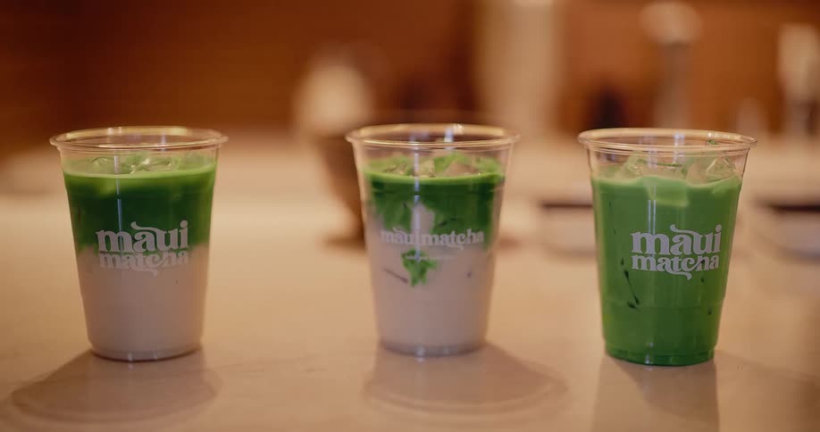 A close-up of three different iced matcha tea lattes in clear cups on a cafe counter