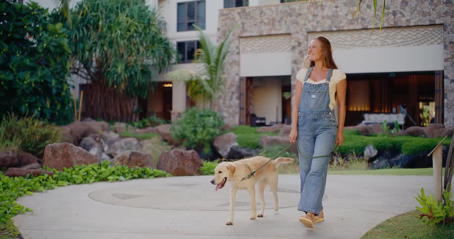 Happy young woman in denim overalls smiles while taking her golden retriever for a leisurely walk through a beautifully landscaped resort