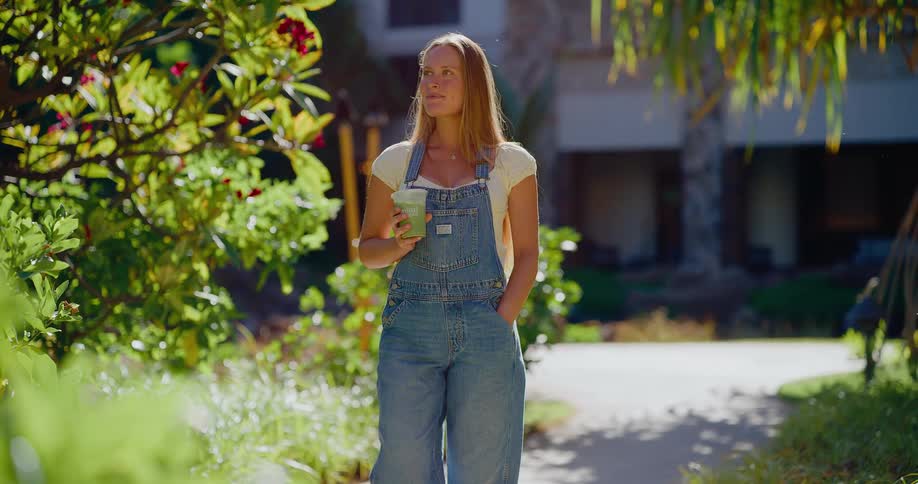 A relaxed young woman in denim overalls holds a green smoothie while standing on a sunlit path in a tropical garden