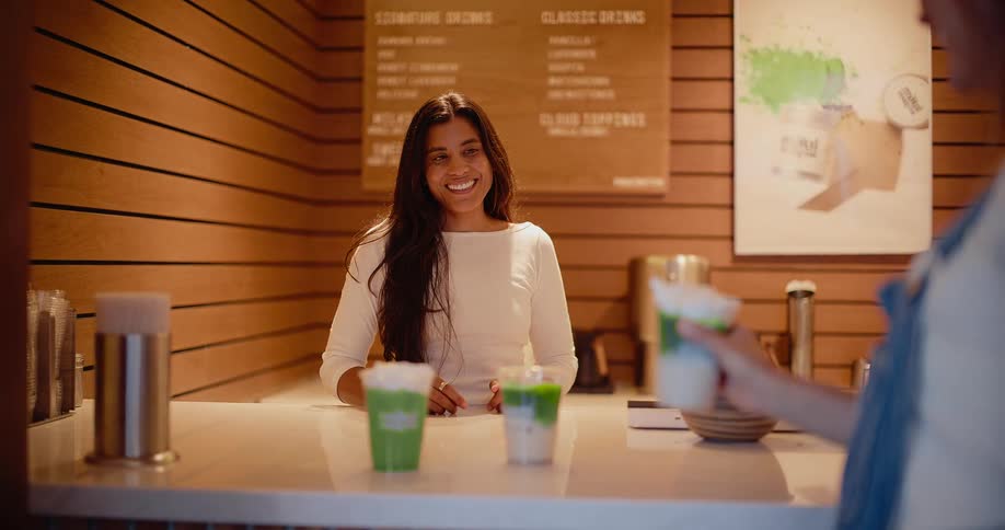 A friendly female barista smiles while serving matcha lattes to a customer at a modern cafe