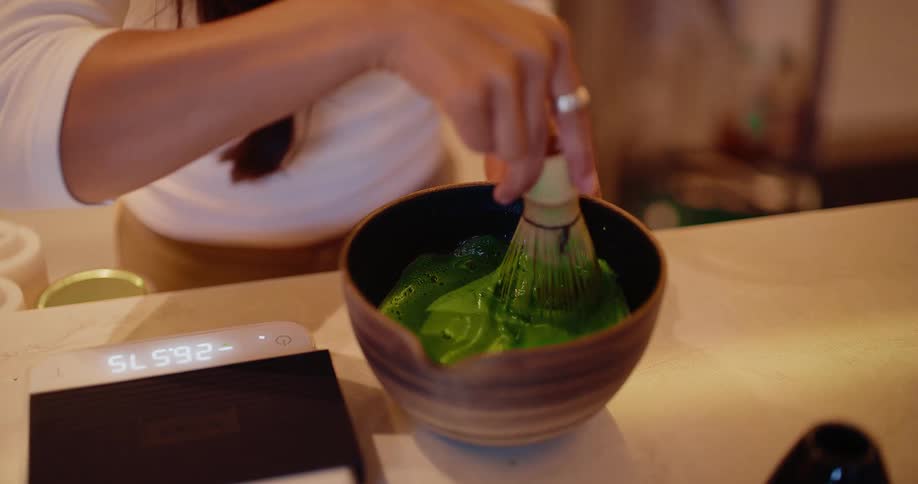 A close-up shot of a person preparing traditional Japanese matcha green tea, whisking it into a frothy consistency in a ceramic bowl