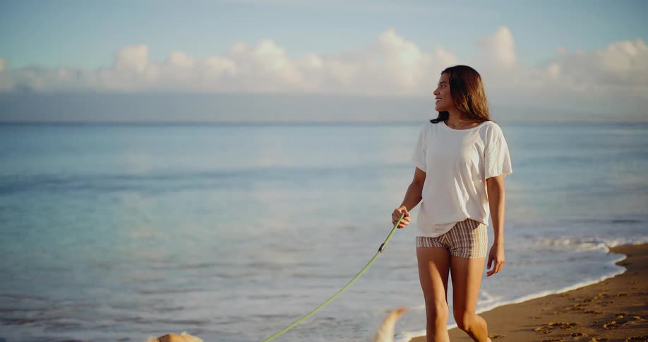 A smiling young woman walks her happy golden retriever on a beautiful beach at sunrise