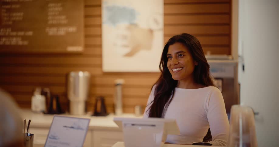 A friendly female cashier with long dark hair smiles brightly while listening to a customer at the counter of a modern cafe