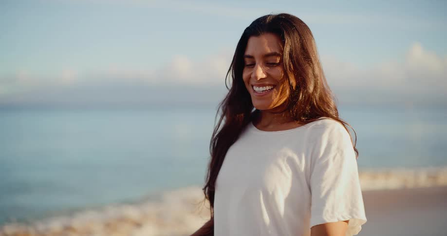 A radiant young woman in a white t-shirt smiles contentedly while enjoying a peaceful moment on a beautiful beach by the ocean