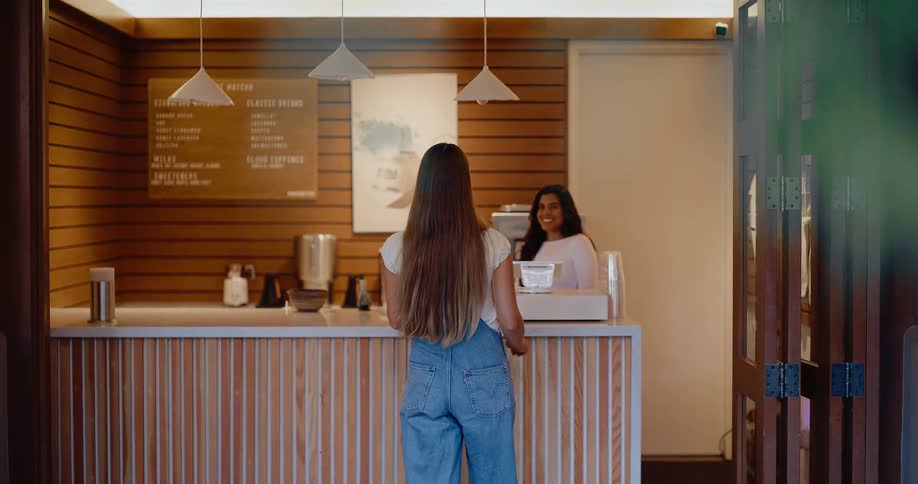 A young woman walks up to a coffee shop counter where a friendly barista waits to take her order