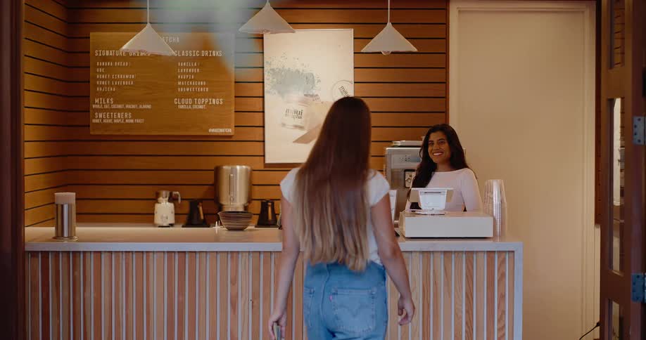 A young woman wearing overalls walks up to the counter of a modern cafe to place her order with a friendly barista