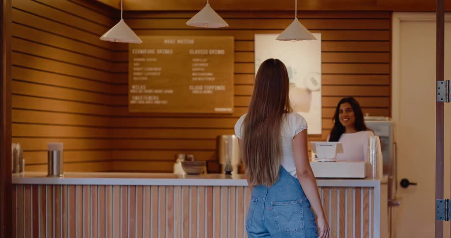 A young woman in overalls approaches the counter to order from a friendly barista in a modern matcha cafe