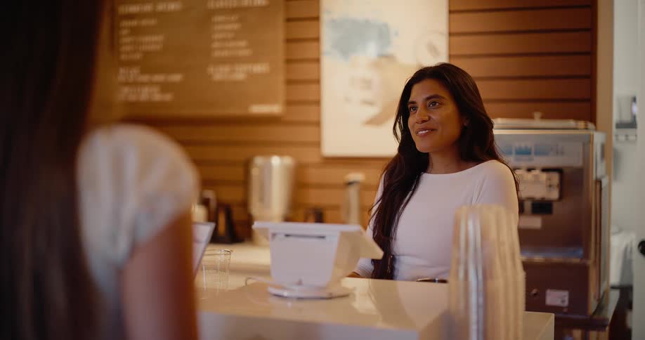 A friendly female barista smiles and listens as she takes a customer's order at a modern coffee shop counter