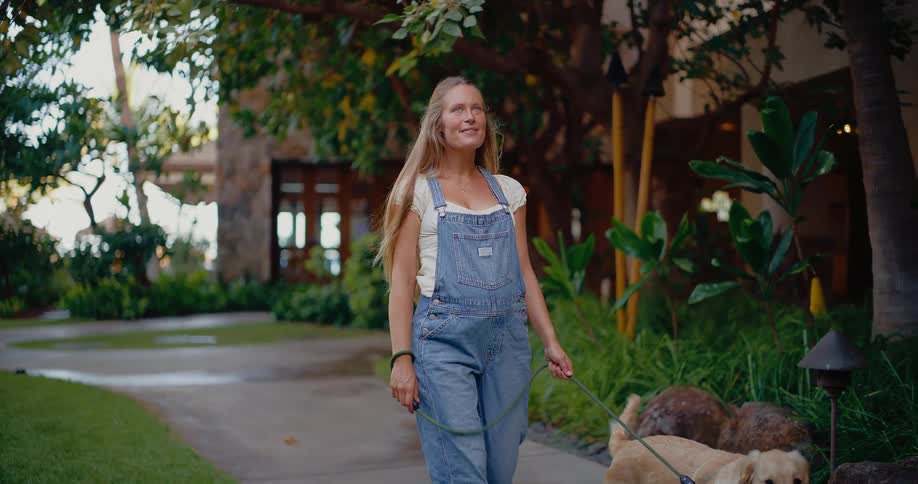 A smiling young woman in denim overalls enjoys a slow-motion walk with her adorable golden retriever on a path through a lush park